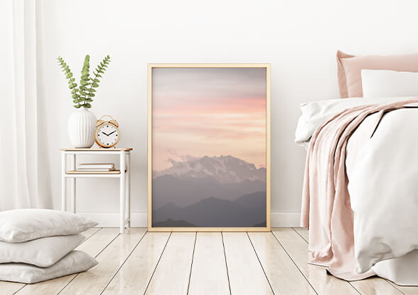 Minimalistic bedroom with light interior and wood floor, showing a bedside table with decorative items on top, a large, photo frame and a bed with white and light pink bedding on the right